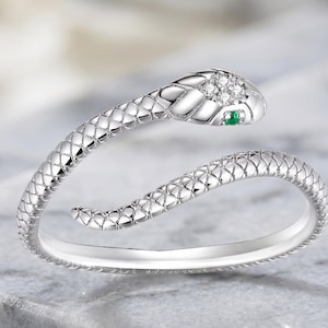 May include: A silver snake ring with a textured, scale-like design. The snake's head features small clear stones and a single green stone for the eye. The ring curves around, creating a unique and elegant look. Jewelry.
