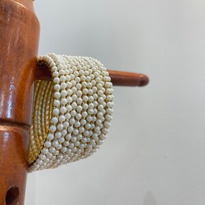 Könnte beinhalten: Ein Stapel goldfarbener Armreifen mit einer dichten, gleichmäßigen Oberfläche aus kleinen, runden, weißen Kunstperlen. Die Armreifen sind zu einer breiten, zylindrischen Manschettenform angeordnet und zeigen ein goldenes Inneres und eine perlenbesetzte Außenseite.