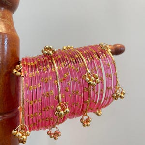 Könnte beinhalten: Ein Set aus durchscheinenden pinkfarbenen Armreifen mit eingearbeiteten goldenen Glitzerpartikeln. Der Stapel enthält mehrere dünne goldfarbene Metallabstandshalter, die jeweils mit kleinen, hängenden goldenen Kugeln verziert sind.
