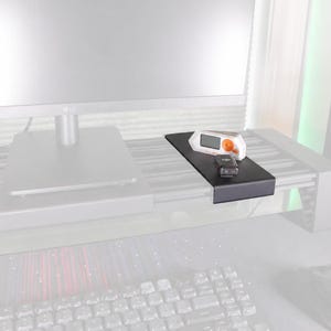 May include: A black monitor shelf with a lip, holding a white and orange device and a black remote. The shelf is mounted on a desk with a keyboard and mouse visible in the foreground. A computer monitor is in the background.