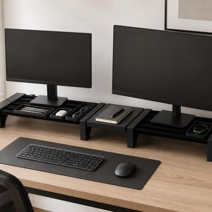 May include: Two black computer monitors sit atop matching black monitor stands with built-in storage compartments. A black keyboard and mouse rest on a black desk mat. A potted plant and a framed picture are visible in the background.
