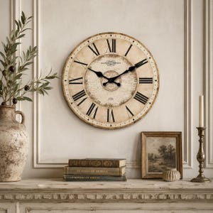 Puede incluir: Un reloj de pared grande y redondo con un diseño vintage. La esfera del reloj es blanquecina con números romanos y manecillas negras. El reloj está rodeado por un marco marrón claro. El reloj está montado en una pared blanca.