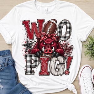 Puede incluir: Una camiseta blanca con un gráfico de un jabalí rojo con un pañuelo, sosteniendo un balón de fútbol americano. El texto WOO PIG! aparece en una mezcla de cuadros rojos, blancos y negros, y texturas de madera, con una estrella negra y un corazón.