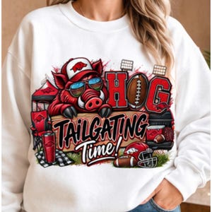 May include: A white crewneck sweatshirt featuring a graphic of a red hog wearing sunglasses and a cap, a football, and the text HOG TAILGATING TIME! The design includes illustrations of a red tent, a red tumbler, and a football helmet on grass.