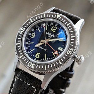 May include: A close-up of a Seiko GMT automatic watch with a black dial and a rotating bezel. The watch has a black leather strap and a silver-colored case. The dial features luminous markers and hands, with the text "SEIKO GMT AUTOMATIC".