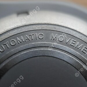 Peut inclure: Gros plan sur un composant de montre avec l'inscription "AUTOMATIC MOVEMENT" grav&eacute;e sur une surface m&eacute;tallique circulaire. La pi&egrave;ce de la montre est de couleur gris clair avec une finition textur&eacute;e, et le texte est en police sans-serif.