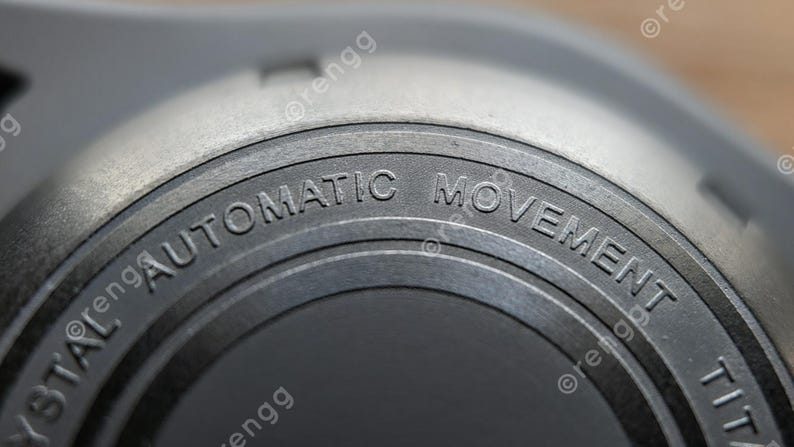 Peut inclure: Gros plan sur un composant de montre avec l'inscription "AUTOMATIC MOVEMENT" grav&eacute;e sur une surface m&eacute;tallique circulaire. La pi&egrave;ce de la montre est de couleur gris clair avec une finition textur&eacute;e, et le texte est en police sans-serif.