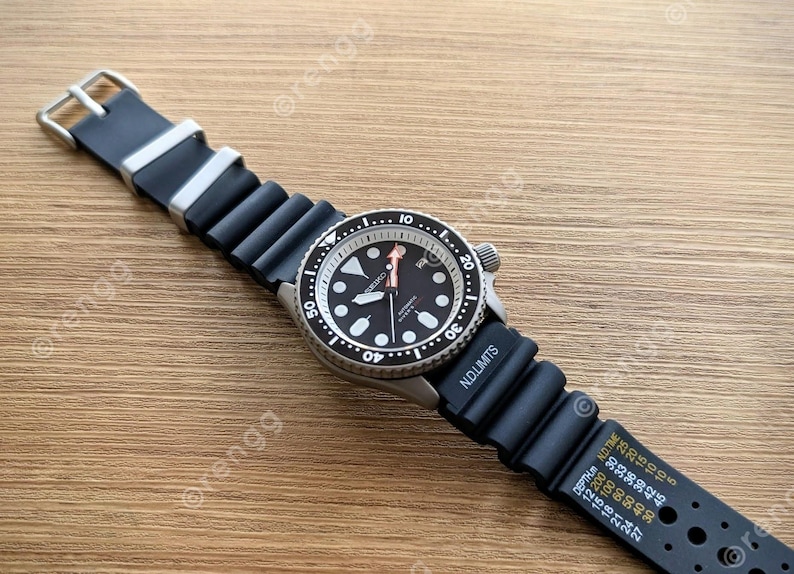 Peut inclure: Une montre-bracelet noire et argent&eacute;e avec un bracelet en caoutchouc noir. Le cadran de la montre est noir avec des marquages et des aiguilles blancs. La montre est sur une surface en bois.
