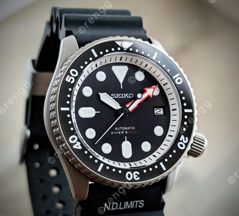 Peut inclure: Gros plan d'une montre de plong&eacute;e automatique Seiko noire avec un cadran noir, des marqueurs blancs et une aiguille des secondes &agrave; pointe rouge. La montre a une lunette en acier inoxydable et un bracelet en caoutchouc noir. Le cadran affiche le texte "SEIKO AUTOMATIC DIVER'S 200m".