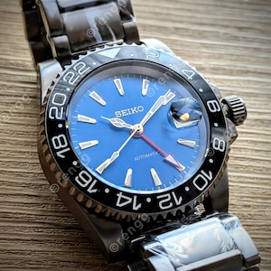 May include: A Seiko automatic wristwatch with a blue dial and a black bezel. The watch features gold-toned hands and markers, a red-tipped second hand, and a stainless steel band. The bezel has white numerals.