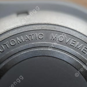 以下が含まれることがあります： 「AUTOMATIC MOVEMENT」の文字が刻まれた、円形の金属表面の時計部品のクローズアップ。時計部品は明るいグレーで、テクスチャード加工が施されており、テキストはサンセリフ体です。