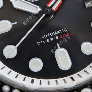 Peut inclure: Gros plan sur un cadran de montre noir avec des marqueurs et des aiguilles blancs. La montre affiche le texte "AUTOMATIC DIVER'S 200m" en blanc et rouge. Elle a une lunette argent&eacute;e avec des marquages blancs.