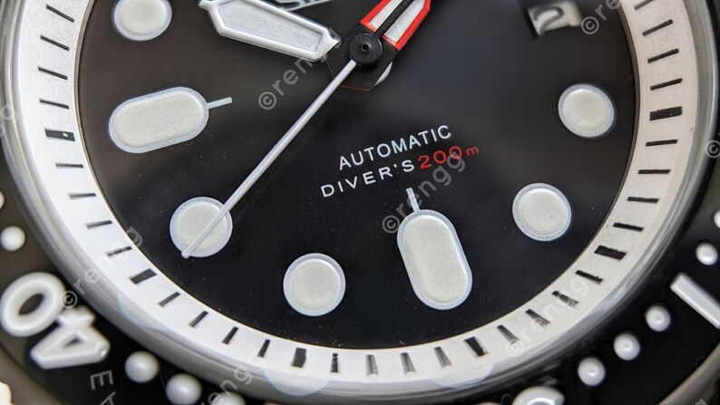 Peut inclure: Gros plan sur un cadran de montre noir avec des marqueurs et des aiguilles blancs. La montre affiche le texte "AUTOMATIC DIVER'S 200m" en blanc et rouge. Elle a une lunette argent&eacute;e avec des marquages blancs.