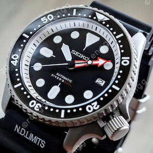 Peut inclure: Une montre de plong&eacute;e automatique Seiko noire avec une lunette noire et un bracelet en caoutchouc noir. Le cadran est noir avec des marqueurs et des aiguilles blancs, et une aiguille des secondes rouge. R&eacute;sistant &agrave; l'eau jusqu'&agrave; 200m.