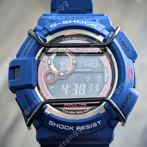 May include: A blue G-Shock digital watch with a pink and black display. The watch features multiple buttons and a black metal protective cage. The display shows the time, date, and day of the week. The watch is labeled "Shock Resist" and "Mud Resist".