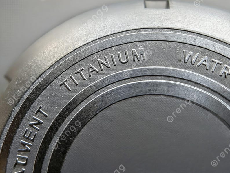 以下が含まれることがあります： 暗い灰色の円形表面に「TITANIUM」の文字がエンボス加工された、金属製の灰色のオブジェクトのクローズアップ。オブジェクトはテクスチャ加工が施されており、より大きなコンポーネントの一部であるように見えます。