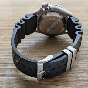 Peut inclure: Gros plan d'une montre-bracelet argent&eacute;e avec un bracelet en caoutchouc noir. La montre a un cadran rond et une lunette textur&eacute;e. Le bracelet noir pr&eacute;sente un motif ondul&eacute; et un fermoir argent&eacute;. La montre est sur une surface en bois.