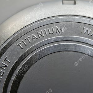 Peut inclure: Gros plan d'un objet gris m&eacute;tallique avec le mot "TITANIUM" en relief sur une surface circulaire gris fonc&eacute;. L'objet a une finition textur&eacute;e et semble faire partie d'un composant plus grand.