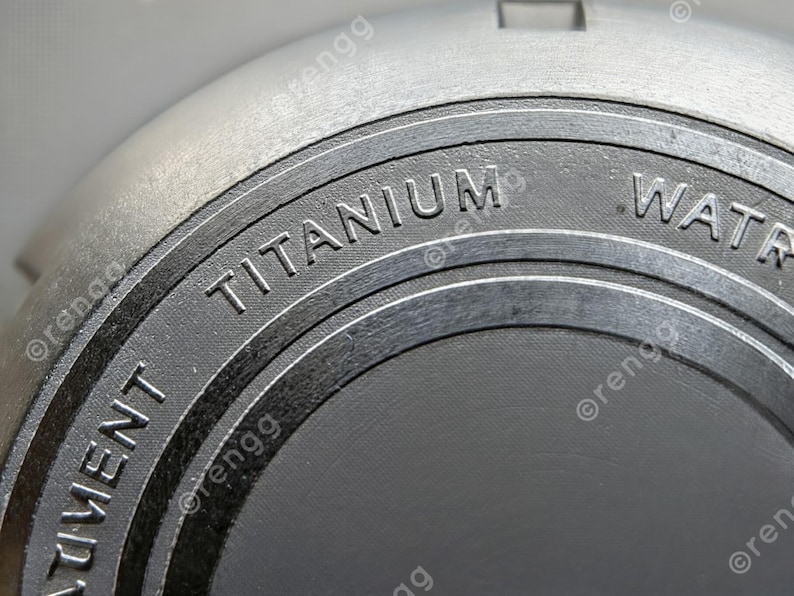 Peut inclure: Gros plan d'un objet gris m&eacute;tallique avec le mot "TITANIUM" en relief sur une surface circulaire gris fonc&eacute;. L'objet a une finition textur&eacute;e et semble faire partie d'un composant plus grand.