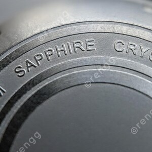 Peut inclure: Gros plan sur un composant de montre avec les mots "SAPPHIRE CRYSTAL" et "200M" grav&eacute;s sur une surface circulaire gris fonc&eacute;. La pi&egrave;ce de la montre a un aspect m&eacute;tallique.