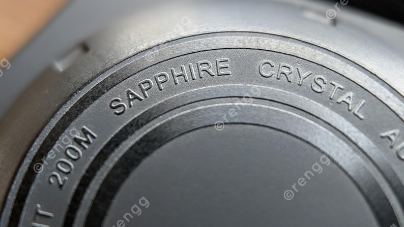 Peut inclure: Gros plan sur un composant de montre avec les mots "SAPPHIRE CRYSTAL" et "200M" grav&eacute;s sur une surface circulaire gris fonc&eacute;. La pi&egrave;ce de la montre a un aspect m&eacute;tallique.