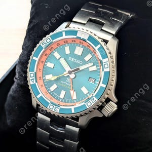 May include: A stainless steel Seiko automatic dive watch with a teal dial and a teal and orange bezel. The watch features a date display and a stainless steel band. The watch is displayed on a black surface.