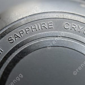 以下が含まれることがあります： 「SAPPHIRE CRYSTAL」と「200M」の文字が刻まれた、濃いグレーの円形表面の時計部品のクローズアップ。 時計部品は金属的な外観をしています。