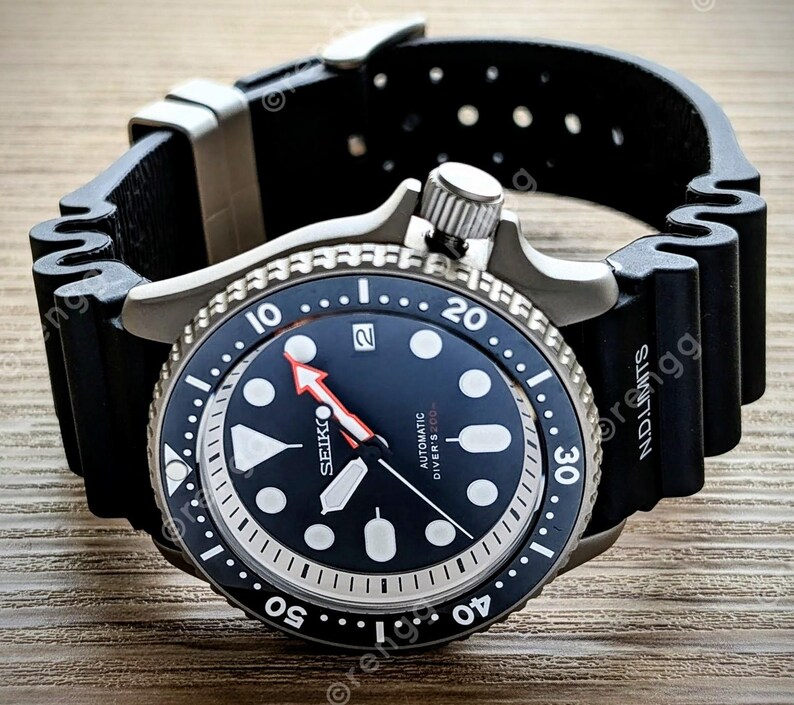 Peut inclure: Une montre de plong&eacute;e automatique Seiko noire et argent&eacute;e avec un bracelet en caoutchouc noir. Le cadran est bleu fonc&eacute; avec des marqueurs blancs et une aiguille des secondes rouge. La lunette a des marquages blancs et le bracelet porte l'inscription "N.D. Limits".