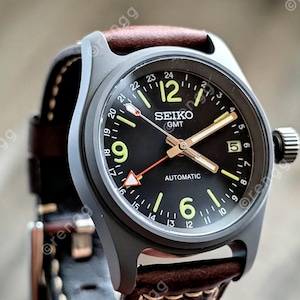 May include: A Seiko GMT automatic watch with a black face, luminous green numerals, and a brown leather strap. The watch has a dark gray case and a date display at the 3 o'clock position. The watch face displays the text "SEIKO GMT AUTOMATIC".