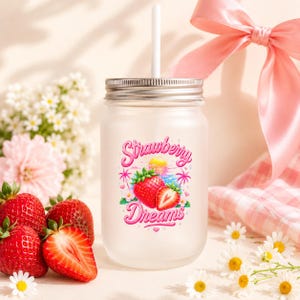 Könnte beinhalten: Ein Trinkglas aus Milchglas in Einmachglasform mit silbernem Deckel und weißem Strohhalm. Auf der Vorderseite befindet sich eine Grafik mit Erdbeeren, Palmen und einem Sonnenuntergang sowie der Schriftzug Strawberry Dreams in rosa Schreibschrift.