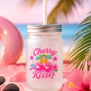 Könnte beinhalten: Ein mattiertes Glas-Einmachglas mit silbernem Metalldeckel und weißem Strohhalm. Die Vorderseite zeigt eine bunte Grafik mit zwei Kirschen, Palmen, einem Sonnenuntergang und einer Hibiskusblüte sowie dem Schriftzug Cherry Kisses in Pink.