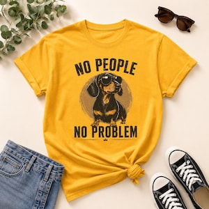 Op de afbeelding: Mosterdgeel T-shirt met een teckel met zonnebril en de tekst "NO PEOPLE NO PROBLEM" in het zwart. De hondenafbeelding is gecentreerd in een cirkelvormig ontwerp. Het shirt wordt gecombineerd met een blauwe jeans en zwarte sneakers.