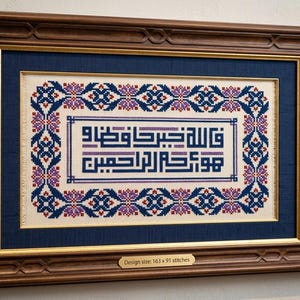 May include: A framed cross-stitch embroidery featuring geometric blue, purple, and red floral borders surrounding Arabic calligraphy in a rectangular format. The dark wood frame includes a small plaque stating Design size: 163 x 91 stitches.
