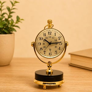 May include: A gold-toned metal desk clock with a circular face mounted on a rotating gimbal frame. The clock features black Arabic numerals for 1-12 and 13-24 hour markings, black hands, and a minute track. It sits on a black cylindrical base.