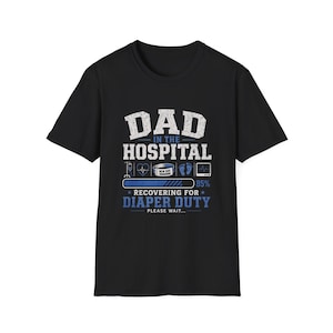 May include: Black t-shirt with a white and blue graphic. The graphic reads "DAD IN THE HOSPITAL" above a loading bar that says "RECOVERING FOR DIAPER DUTY". The shirt also features medical icons and the text "PLEASE WAIT..."