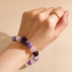 May include: A beaded bracelet featuring a mix of purple, pink, and clear gemstones. The bracelet includes round and square beads. A gold ring is on the finger. The bracelet is on a wrist.