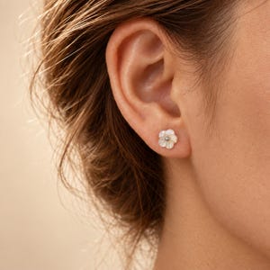 Könnte beinhalten: Ein kleiner Ohrstecker in Form einer fünfblättrigen Blume. Die Blütenblätter bestehen aus irisierendem, weißem Perlmutt mit einer glatten, glänzenden Oberfläche. In der Mitte der Blume befindet sich eine winzige, polierte silberfarbene Metallperle.