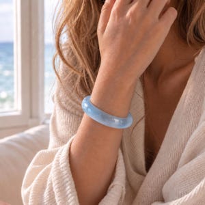 May include: A smooth, solid light blue bangle bracelet with a polished, translucent finish. The circular accessory features a rounded profile and a consistent thickness, showcasing a clear, glass-like appearance that catches the light.