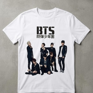 Pode incluir: Camiseta branca com o texto preto "BTS" e um grupo de sete pessoas vestidas de preto. A camiseta tem gola redonda e mangas curtas. O texto abaixo do logotipo BTS está em outro idioma.