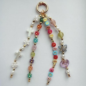 May include: A beaded keyring featuring a gold-toned metal ring with three dangling strands. The strands include white cross beads, colourful flower-shaped beads, a strand spelling JESUS LOVES YOU, and a butterfly and heart-shaped charm.