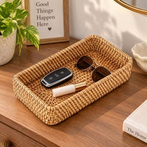 May include: A rectangular woven rattan tray with a light brown natural finish. Inside the tray, there is a black car key fob with silver buttons, a pair of brown-tinted sunglasses with thin gold-tone frames, and a small white cylindrical tube.
