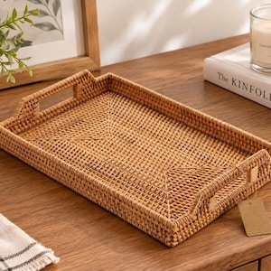 May include: A rectangular woven rattan tray featuring a light brown natural finish, a flat base, and two integrated handles on the shorter sides. A small, blank, rectangular beige paper tag is attached to one of the handles with a thin string.