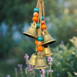 May include: A cluster of small, antique brass bells hanging from a natural rope. The rope is adorned with colorful wooden beads in shades of teal, orange, and brown. The bells are suspended against a blurred background of green foliage and purple flowers.