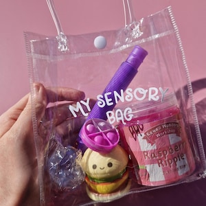 Puede incluir: Un bolso de plástico transparente con el texto blanco MY SENSORY BAG, que contiene un tubo pop morado, un pequeño juguete fidget circular rosa, un juguete squishy con forma de hamburguesa y un bote de slime perfumado Raspberry Ripple.