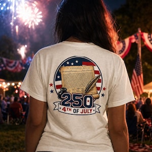 May include: White t-shirt featuring a design commemorating the 250th anniversary of the 4th of July. The design includes a scroll, quill, inkwell, and stars within a circular emblem. Fireworks and an American flag are visible in the background.