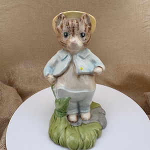 May include: A ceramic figurine of a tabby cat wearing a light blue jacket and a yellow hat. The cat stands on a green grassy base with a small gray stone detail. The figure features brown stripes on its head and a white shirt underneath the jacket.
