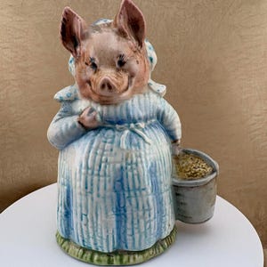 May include: A ceramic figurine of a pig dressed in a blue and white checkered dress and a matching bonnet. The pig has a pinkish-brown complexion and is holding a small, textured gray bucket in its left hand while standing on a green base.