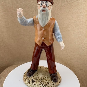 Może przedstawiać: Ceramiczna figurka starszej osoby z długą białą brodą, ubranej w brązową czapkę z daszkiem, jasnoniebieską koszulę, beżową kamizelkę i ciemnobrązowe spodnie. Postać ma okulary, otwarte usta i uniesioną prawą pięść, stojąc na podstawie.
