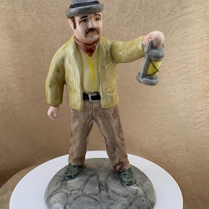 May include: A ceramic figurine of a person wearing a grey hat, a yellow jacket, and brown trousers. The figure has a mustache and is holding a small grey lantern in one hand. The figurine stands on a circular base with a grey, textured stone-like pattern.