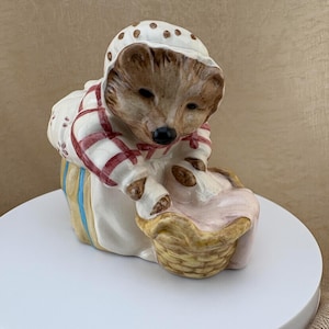 Peut inclure: Une figurine de hérisson en céramique portant un bonnet blanc à pois marron, une chemise blanche aux manches à carreaux rouges et un tablier rayé jaune et bleu. Le hérisson est penché en avant, atteignant un petit panier en osier beige contenant du tissu rose.